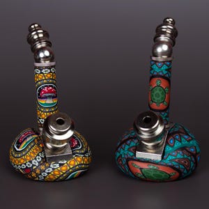 May include: Two smoking pipes with metallic stems and bowls. The left pipe features a yellow and black patterned base with a red mushroom design. The right pipe has a teal and orange patterned base with a green turtle design on the body.