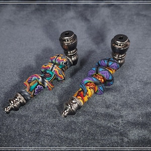 May include: Two decorative pipes with colorful, patterned designs. One pipe features a teal, purple, and yellow design, while the other showcases a purple, orange, and yellow pattern. Both pipes have silver metal accents and dark brown bowls.
