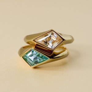 Könnte beinhalten: Zwei goldene Ringe mit geometrischen, rautenförmigen Edelsteinen. Ein Ring hat einen klaren, facettierten Stein, der andere einen hellblauen Edelstein. Die Ringe sind gestapelt, der blaue Ring im Vordergrund.