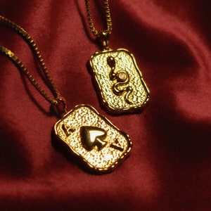 Könnte beinhalten: Zwei goldene Halsketten mit Anhänger-Anhängern. Ein Anhänger ist eine Schlange mit einem rosa Stein, der andere ist ein Pik-Ass.