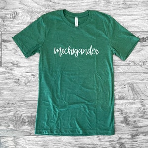 Puede incluir: Una camiseta verde con la palabra "Michigander" en escritura blanca en la parte delantera. La camiseta tiene mangas cortas y cuello redondo. La camiseta est&aacute; hecha de un material suave. Se muestra sobre una superficie de madera clara.