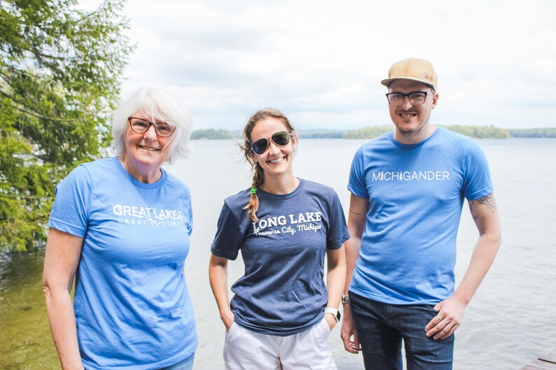 Puede incluir: Tres personas con camisetas con texto. Una camiseta dice "GREAT LAKES GREAT TIMES" en blanco, otra dice "LONG LAKE Traverse City, Michigan" en blanco, y la tercera dice "MICHIGANDER" en blanco. Est&aacute;n de pie cerca de un lago.