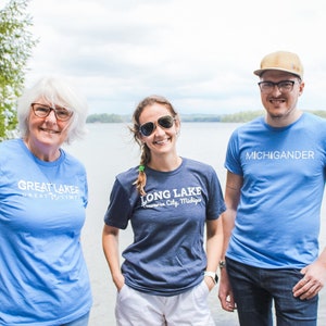 Może przedstawiać: Trzy osoby w koszulkach z tekstem. Jedna koszulka ma napis "GREAT LAKES GREAT TIMES" w kolorze białym, inna "LONG LAKE Traverse City, Michigan" w kolorze białym, a trzecia "MICHIGANDER" w kolorze białym. Stoją nad jeziorem.