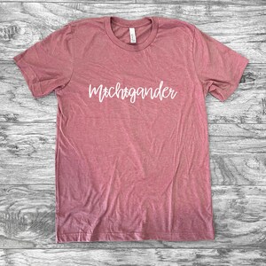 Puede incluir: Una camiseta de color rosa polvoriento con la palabra "Michigander" en escritura blanca. La camiseta de manga corta est&aacute; hecha de un material suave y est&aacute; colocada sobre una superficie de madera. La camiseta tiene cuello redondo.