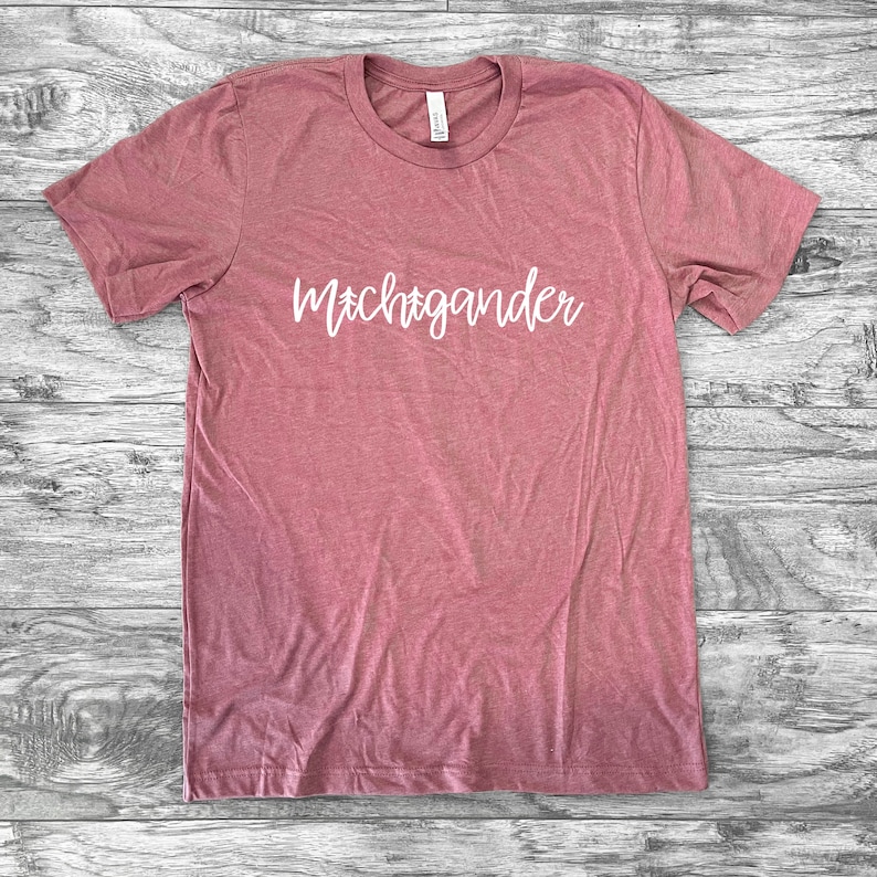 Puede incluir: Una camiseta de color rosa polvoriento con la palabra "Michigander" en escritura blanca. La camiseta de manga corta est&aacute; hecha de un material suave y est&aacute; colocada sobre una superficie de madera. La camiseta tiene cuello redondo.