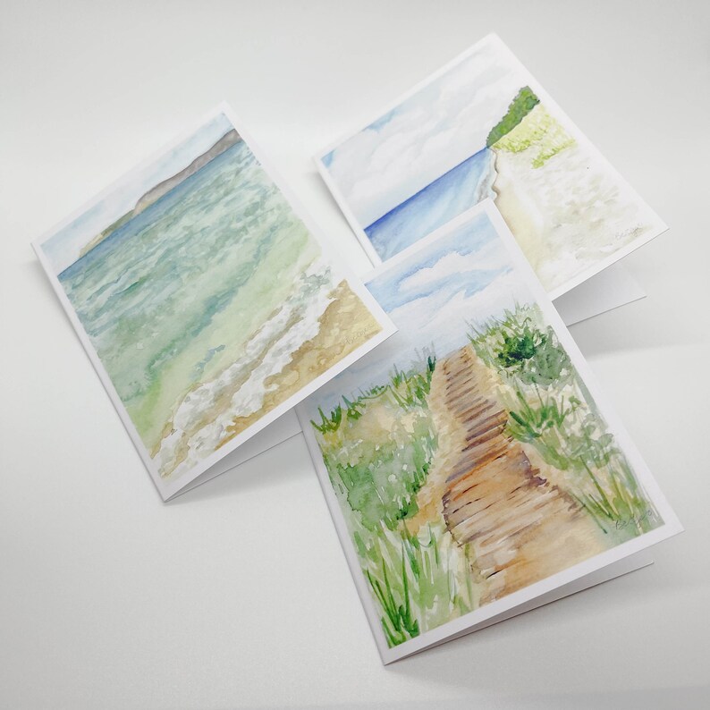 Puede incluir: Juego de tres tarjetas de felicitaci&oacute;n de acuarela con escenas de playa. Cada tarjeta representa una vista costera diferente: olas del oc&eacute;ano, una playa con dunas y un paseo mar&iacute;timo de madera a trav&eacute;s de hierba alta. Las tarjetas est&aacute;n en blanco por dentro.