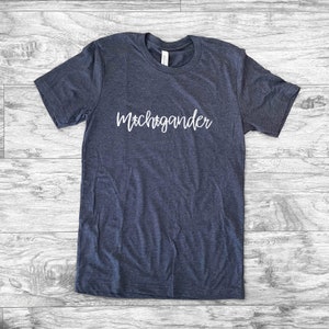 Puede incluir: Una camiseta azul marino con la palabra "Michigander" en escritura blanca en el pecho. La camiseta est&aacute; hecha de un material suave y tiene mangas cortas. La camiseta est&aacute; tendida sobre una superficie de madera.