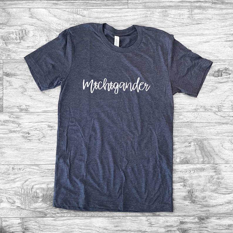 Puede incluir: Una camiseta azul marino con la palabra "Michigander" en escritura blanca en el pecho. La camiseta est&aacute; hecha de un material suave y tiene mangas cortas. La camiseta est&aacute; tendida sobre una superficie de madera.