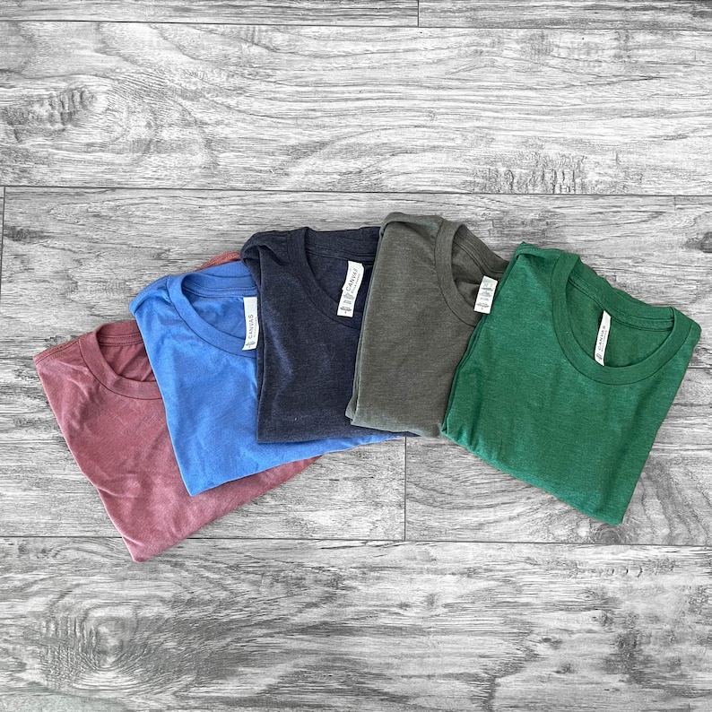 Puede incluir: Cinco camisetas de cuello redondo de colores lisos, dispuestas en fila. Los colores son rosa empolvado, azul claro, azul marino, verde oliva y verde bosque. Cada camiseta tiene una etiqueta en el cuello.