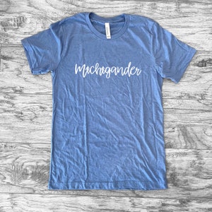 Puede incluir: Una camiseta azul claro con la palabra "Michigander" en escritura blanca en el pecho. La camiseta es de un material suave y tiene cuello redondo. La camiseta est&aacute; tendida sobre una superficie de madera.