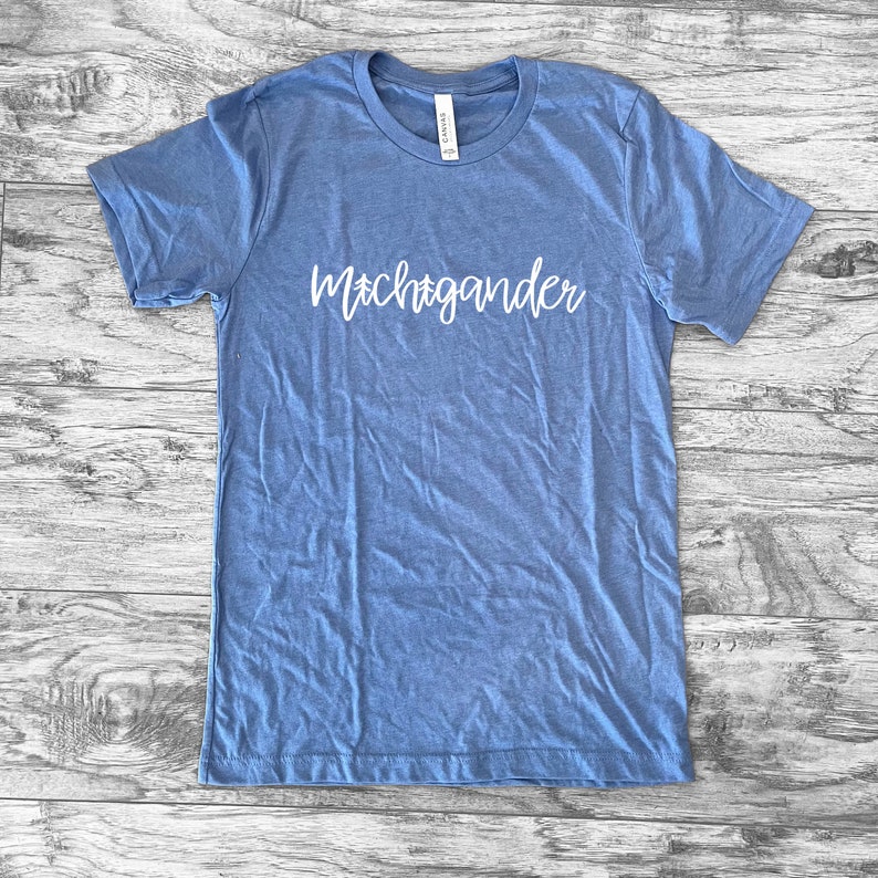 Puede incluir: Una camiseta azul claro con la palabra "Michigander" en escritura blanca en el pecho. La camiseta es de un material suave y tiene cuello redondo. La camiseta est&aacute; tendida sobre una superficie de madera.