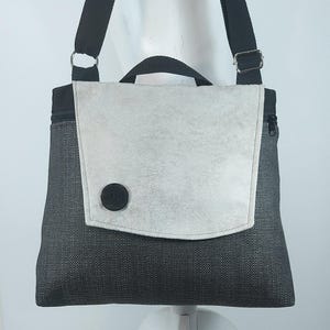 Pode incluir: Bolsa transversal cinza e preta com fecho de aba. A bolsa apresenta um corpo de tecido cinza texturizado, uma aba cinza claro presa por um botão preto e uma alça de ombro preta ajustável. Uma alça preta também é visível.