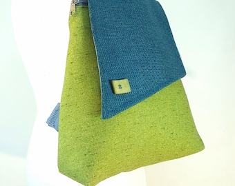 May include: A handmade backpack with a textured green body and a blue flap. The backpack has a top handle and a zipper closure. A small green square with a button is on the flap. The backpack is displayed on a white mannequin.