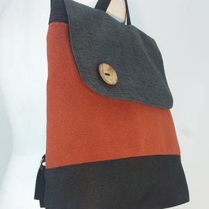 May include: A backpack with a flap closure, featuring a color-blocked design. The top flap is gray, the middle section is orange, and the bottom is black. A wooden button secures the flap. The bag has black straps.