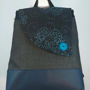 May include: A backpack with a dark gray woven fabric body and a navy blue faux leather base. The flap is black with a teal swirl pattern and a teal button closure. A black handle and zipper are visible.