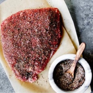 May include: A raw piece of meat seasoned with black pepper and salt on a piece of parchment paper.