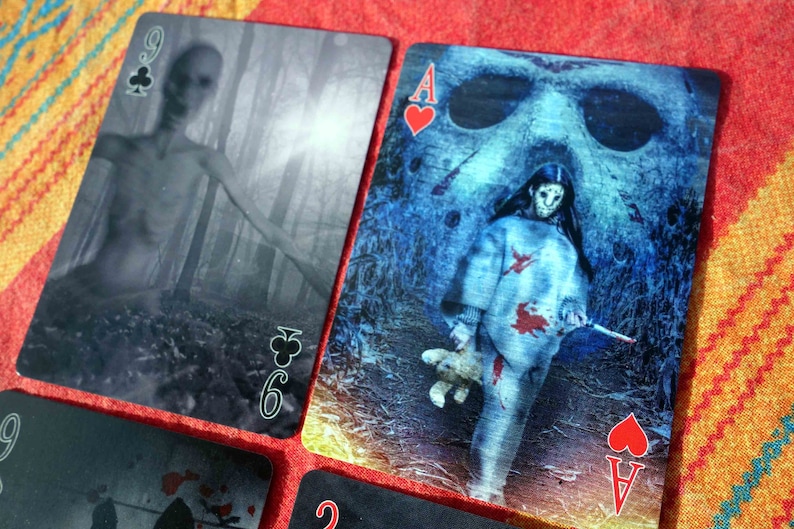 Puede incluir: Dos cartas de juego, un 9 de tr&eacute;boles y un as de corazones, con ilustraciones de tem&aacute;tica de pel&iacute;culas de terror. El 9 de tr&eacute;boles presenta una imagen borrosa de una figura en un bosque. El as de corazones presenta una figura con una m&aacute;scara de hockey y sosteniendo un cuchillo ensangrentado.