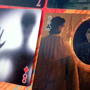 Puede incluir: Dos cartas de juego, una es el 8 de diamantes con una imagen borrosa de una mano y una figura, la otra es el 2 de tr&eacute;boles con una persona mirando en un espejo con una figura aterradora reflejada en el espejo.
