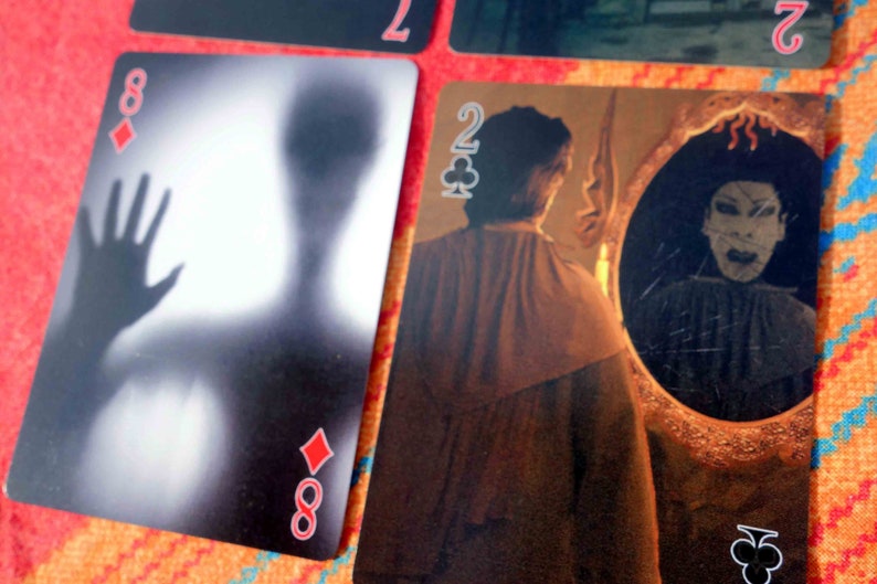 Puede incluir: Dos cartas de juego, una es el 8 de diamantes con una imagen borrosa de una mano y una figura, la otra es el 2 de tr&eacute;boles con una persona mirando en un espejo con una figura aterradora reflejada en el espejo.