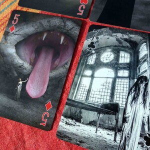 Puede incluir: Dos cartas de juego, un 5 de diamantes y un 7 de tr&eacute;boles, con un dise&ntilde;o oscuro y g&oacute;tico. El 5 de diamantes presenta una gran boca abierta con una larga lengua rosa. El 7 de tr&eacute;boles muestra a una mujer con un vestido blanco de pie en un edificio en ruinas con una gran ventana.