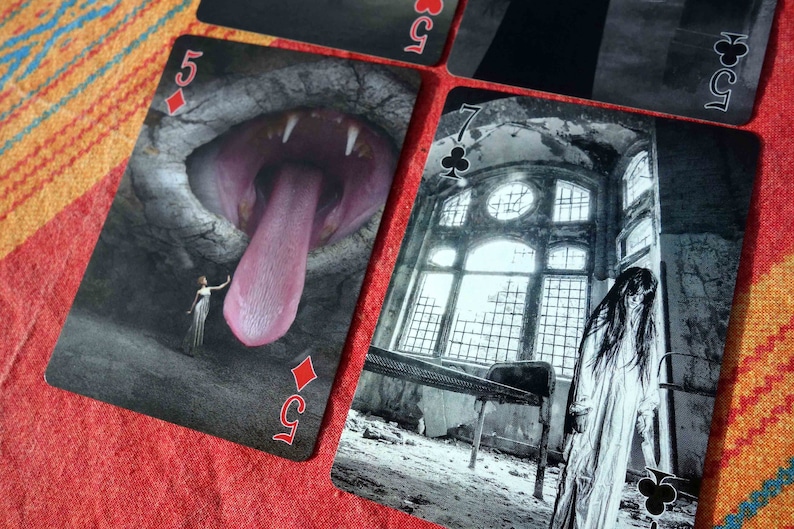 Puede incluir: Dos cartas de juego, un 5 de diamantes y un 7 de tr&eacute;boles, con un dise&ntilde;o oscuro y g&oacute;tico. El 5 de diamantes presenta una gran boca abierta con una larga lengua rosa. El 7 de tr&eacute;boles muestra a una mujer con un vestido blanco de pie en un edificio en ruinas con una gran ventana.