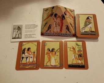 May include: A set of Oracle of Pyramids cards with illustrations of Egyptian figures and symbols. The cards are in a pyramid shape with the text "Oracle of Pyramids" on the front. The cards are on a light brown surface.