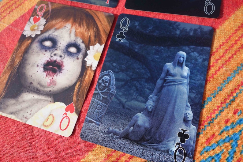 Puede incluir: Dos cartas de juego, ambas son reinas. La reina de corazones presenta una mu&ntilde;eca espeluznante con cabello rojo y una boca ensangrentada. La reina de tr&eacute;boles presenta una escena azul y sombr&iacute;a con una mujer con un vestido largo de pie sobre otras dos figuras.