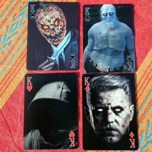Puede incluir: Cuatro cartas de juego con diferentes reyes. La primera carta muestra un rey con una cabeza de calabaza sosteniendo un cuchillo. La segunda carta muestra un rey con un cuerpo azul y una corona. La tercera carta muestra un rey con una figura negra encapuchada. La cuarta carta muestra un rey con una cara que tiene una expresi&oacute;n seria.