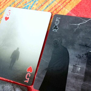 Puede incluir: Dos cartas de juego, un cinco de corazones y un cinco de tr&eacute;boles, con un dise&ntilde;o oscuro, brumoso y espeluznante. La carta cinco de corazones muestra una figura caminando en la niebla, mientras que la carta cinco de tr&eacute;boles muestra una figura de pie en un cementerio.