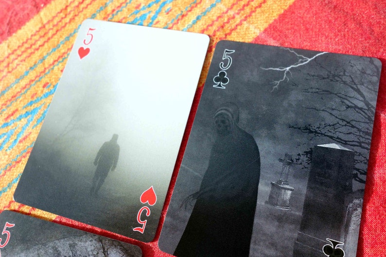 Puede incluir: Dos cartas de juego, un cinco de corazones y un cinco de tr&eacute;boles, con un dise&ntilde;o oscuro, brumoso y espeluznante. La carta cinco de corazones muestra una figura caminando en la niebla, mientras que la carta cinco de tr&eacute;boles muestra una figura de pie en un cementerio.