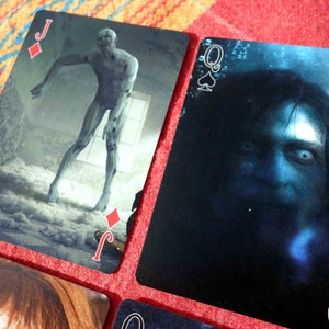 Puede incluir: Cuatro cartas de juego de una baraja de tem&aacute;tica de terror. Las cartas son el Jack de Diamantes, la Reina de Espadas, la Reina de Corazones y la Reina de Tr&eacute;boles. Cada carta presenta una imagen aterradora diferente.