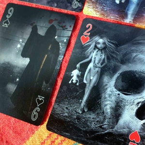 Puede incluir: Dos cartas de juego, un 9 de picas y un 2 de corazones, con im&aacute;genes oscuras y g&oacute;ticas. El 9 de picas presenta una figura sombr&iacute;a con una guada&ntilde;a, mientras que el 2 de corazones representa a una mujer con una calavera y un animal peque&ntilde;o.