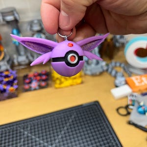 May include: A purple and black 3D printed keychain featuring a Pokemon character. The character has large, pointed ears and a round body with a red and black Pokeball design.