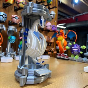 Puede incluir: Una figura de Pok&eacute;mon impresa en 3D de color blanco de un Pok&eacute;mon p&aacute;jaro legendario, con detalles azules, de pie sobre un pedestal plateado. La figura est&aacute; en una pose con las alas extendidas.