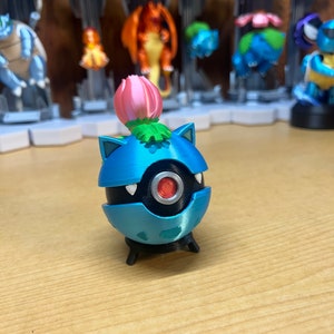 Puede incluir: Una figura de Pok&eacute;mon impresa en 3D de color azul y negro con una flor rosa en la parte superior. La figura tiene un ojo rojo y una base negra.