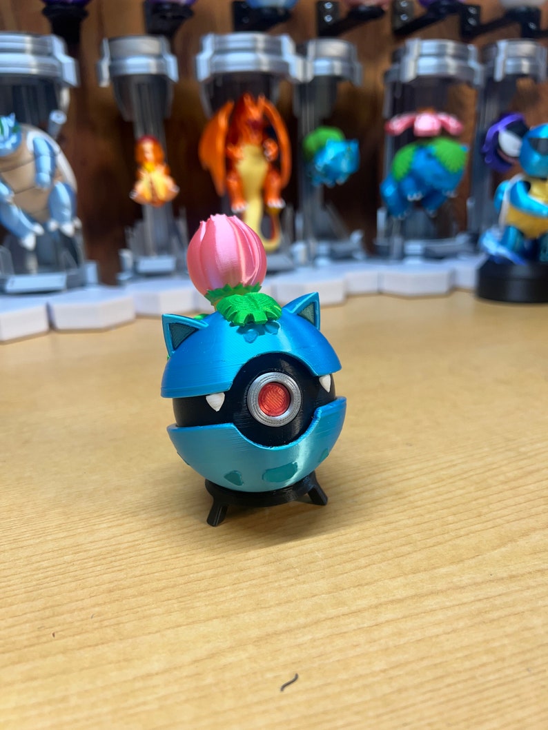 Puede incluir: Una figura de Pok&eacute;mon impresa en 3D de color azul y negro con una flor rosa en la parte superior. La figura tiene un ojo rojo y una base negra.
