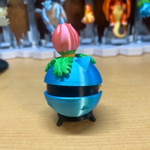 Puede incluir: Un juguete de impresi&oacute;n 3D azul y negro con una flor rosa en la parte superior. El juguete tiene forma de esfera con una peque&ntilde;a base.