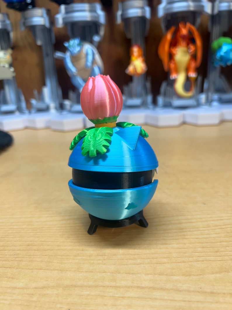 Puede incluir: Un juguete de impresi&oacute;n 3D azul y negro con una flor rosa en la parte superior. El juguete tiene forma de esfera con una peque&ntilde;a base.