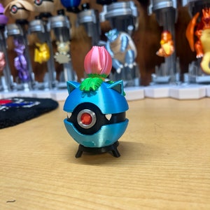 Puede incluir: Una figura de Pok&eacute;mon impresa en 3D de color azul y negro con una flor rosa en la parte superior. La figura est&aacute; de pie sobre una base negra.