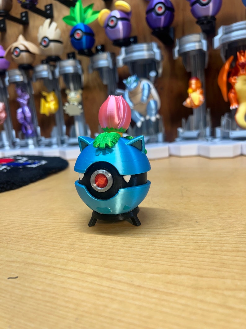 Puede incluir: Una figura de Pok&eacute;mon impresa en 3D de color azul y negro con una flor rosa en la parte superior. La figura est&aacute; de pie sobre una base negra.