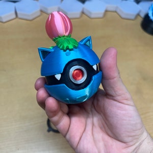 Puede incluir: Una figura impresa en 3D de color azul y negro de un personaje de Pokemon con una flor rosa en la parte superior. El personaje tiene un cuerpo redondo con un ojo negro y una boca blanca.