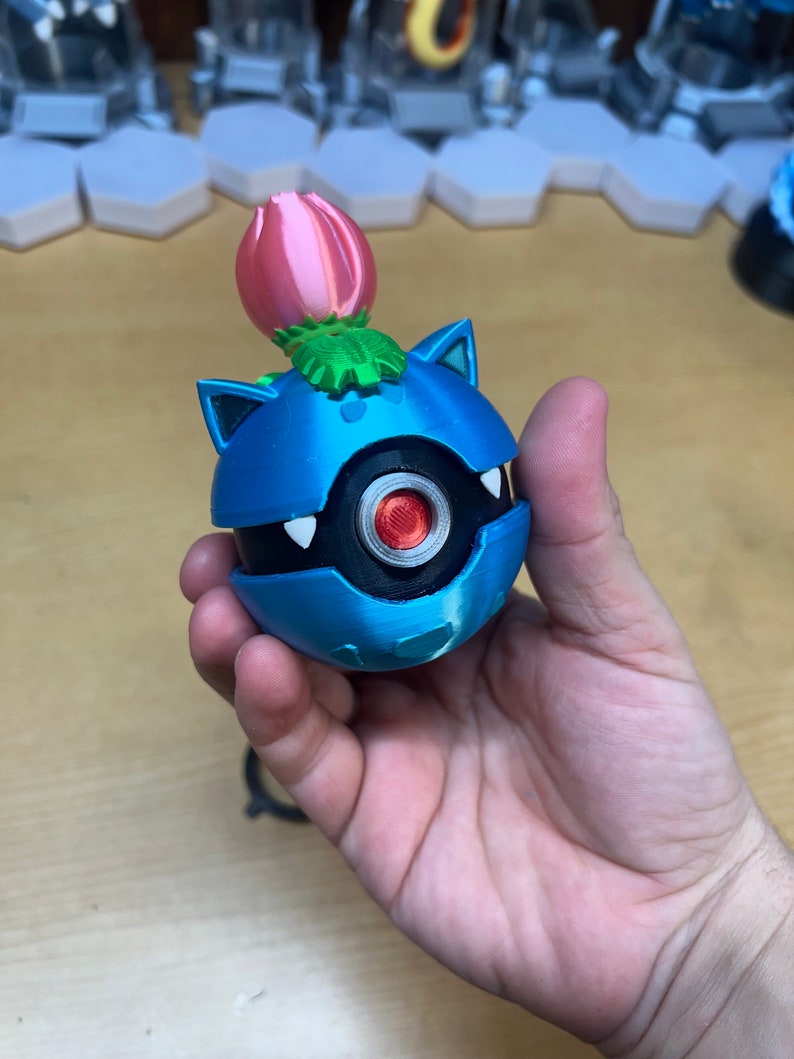 Puede incluir: Una figura impresa en 3D de color azul y negro de un personaje de Pokemon con una flor rosa en la parte superior. El personaje tiene un cuerpo redondo con un ojo negro y una boca blanca.