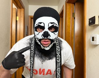 May include: A black knit balaclava featuring a white skull graphic print, worn with a white t-shirt displaying red text and a long black strap with repeating white text. The attire includes black gloves and black pants.
