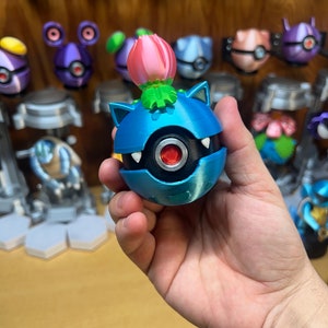 Puede incluir: Una figura Pok&eacute;mon impresa en 3D de color azul y plateado con una flor rosa en la parte superior. La figura est&aacute; siendo sostenida en la mano de una persona.