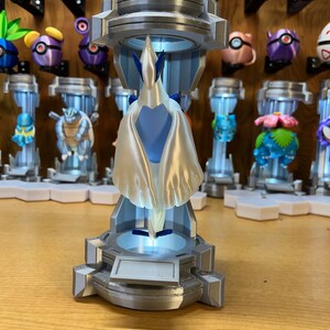 Puede incluir: Un modelo 3D impreso de un personaje Pok&eacute;mon blanco y azul, de pie sobre una base plateada con una c&uacute;pula de pl&aacute;stico transparente. El personaje est&aacute; en pose con las alas extendidas.