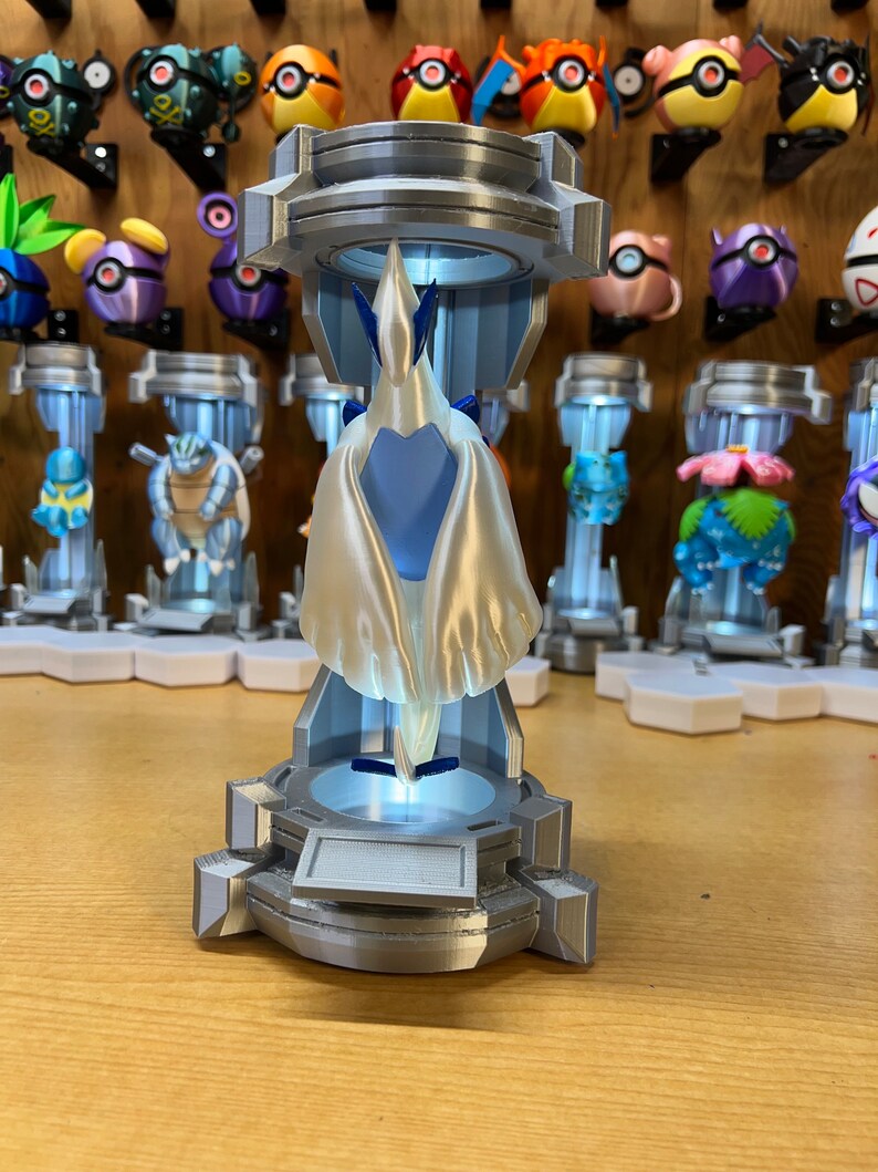Puede incluir: Un modelo 3D impreso de un personaje Pok&eacute;mon blanco y azul, de pie sobre una base plateada con una c&uacute;pula de pl&aacute;stico transparente. El personaje est&aacute; en pose con las alas extendidas.