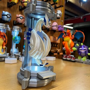 Puede incluir: Una figura blanca de un personaje Pok&eacute;mon, Lugia, impresa en 3D, en una vitrina de pl&aacute;stico transparente con una base de luz azul. La figura est&aacute; posada con las alas extendidas y la cabeza inclinada hacia un lado. La base tiene un esquema de color plateado y gris.