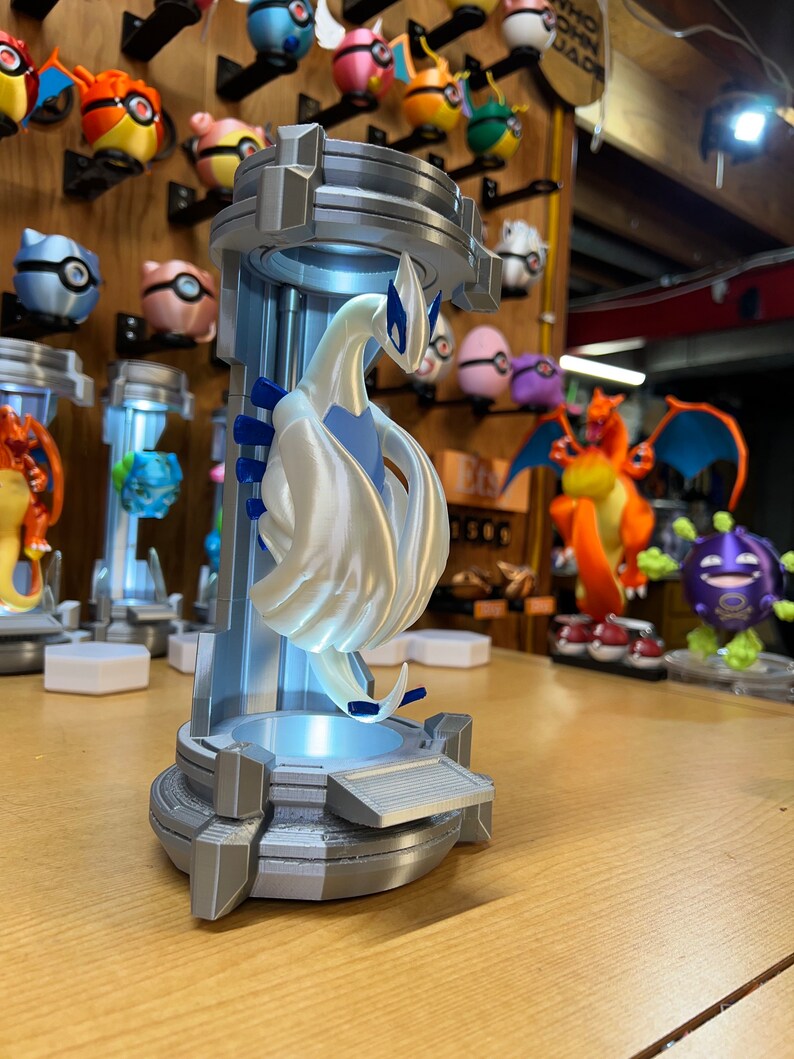 Puede incluir: Una figura blanca de un personaje Pok&eacute;mon, Lugia, impresa en 3D, en una vitrina de pl&aacute;stico transparente con una base de luz azul. La figura est&aacute; posada con las alas extendidas y la cabeza inclinada hacia un lado. La base tiene un esquema de color plateado y gris.