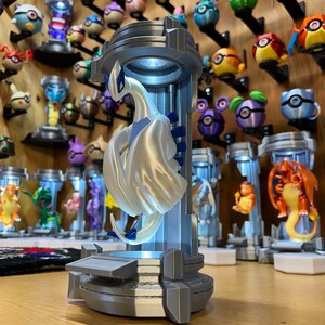 Puede incluir: Una figura impresa en 3D de color blanco y azul de una criatura m&iacute;tica, que se asemeja a un drag&oacute;n, se exhibe en un cilindro de pl&aacute;stico transparente con una base gris. La figura est&aacute; iluminada desde el interior, proyectando un suave resplandor. La figura est&aacute; rodeada de otras figuras similares, todas expuestas sobre una superficie de madera.