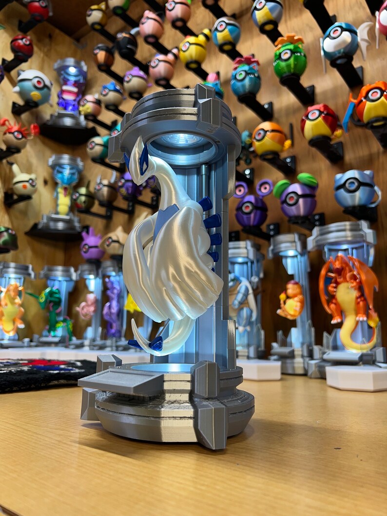 Puede incluir: Una figura impresa en 3D de color blanco y azul de una criatura m&iacute;tica, que se asemeja a un drag&oacute;n, se exhibe en un cilindro de pl&aacute;stico transparente con una base gris. La figura est&aacute; iluminada desde el interior, proyectando un suave resplandor. La figura est&aacute; rodeada de otras figuras similares, todas expuestas sobre una superficie de madera.